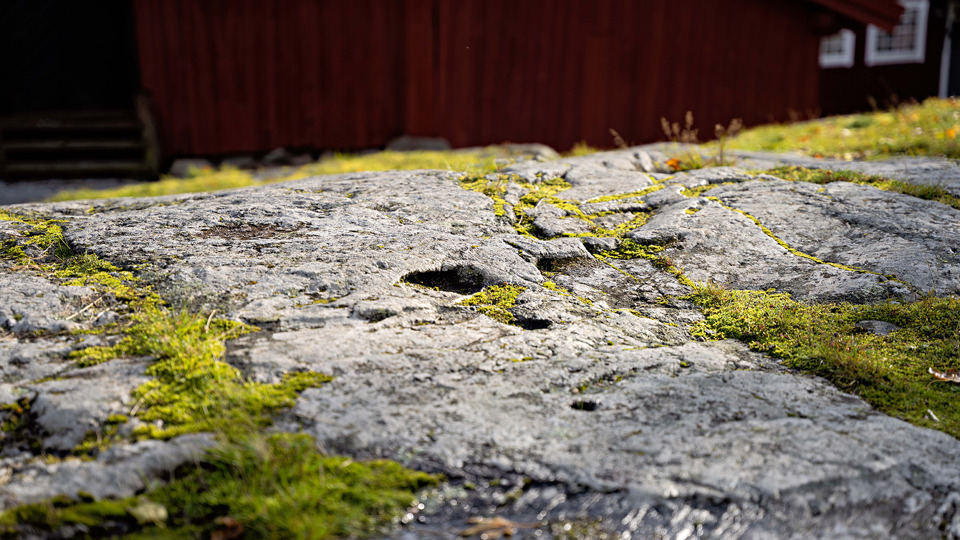 Stenhäll med mossa. I bakgrunden syns en röd träbyggnad.