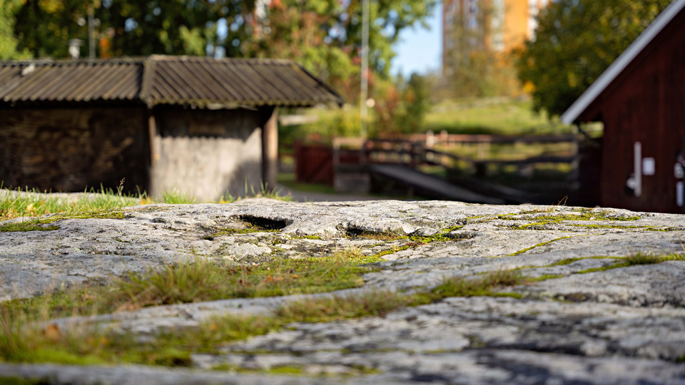 Stenhäll med mossa. I bakgrunden syns gångväg, staket och två mindre hus.