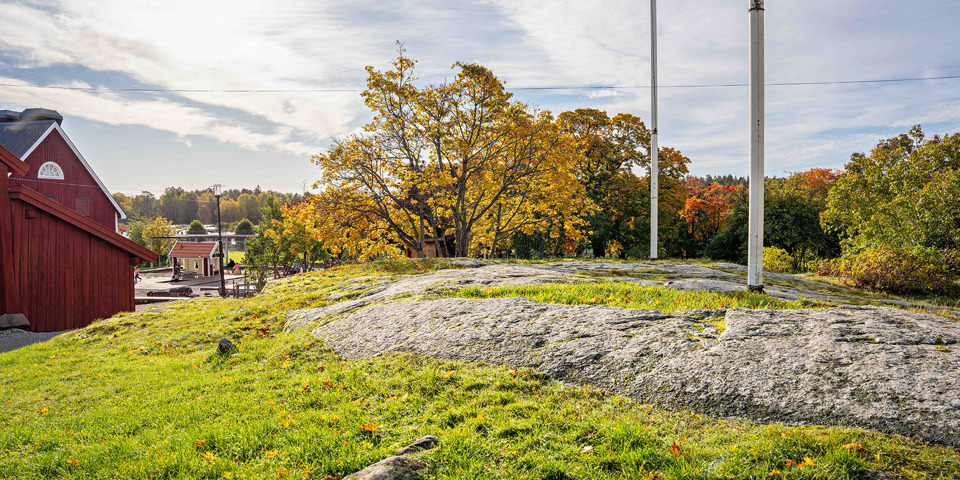 Kulle med stenhäll och gräs. Bredvid finns träd och en röd byggnad.